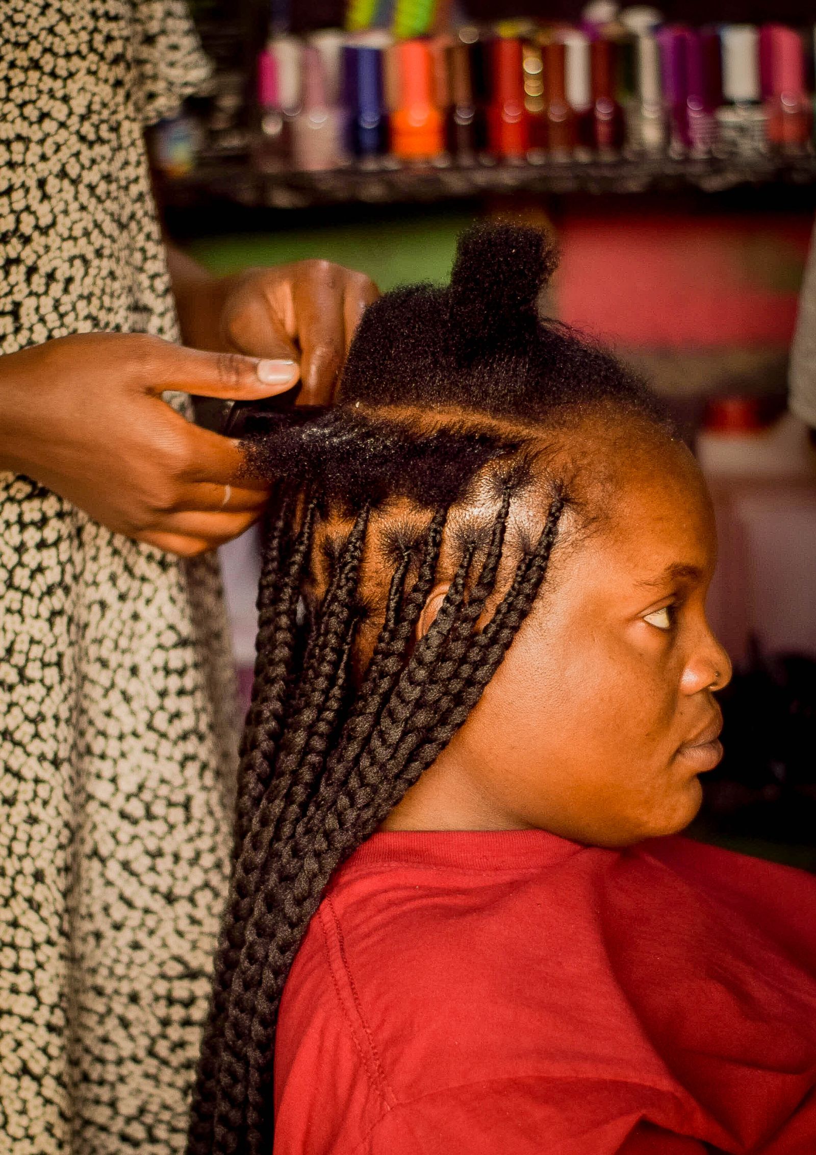 A Lagos stylist braiding long knotless braids into a seated client's hair, clean parts visible