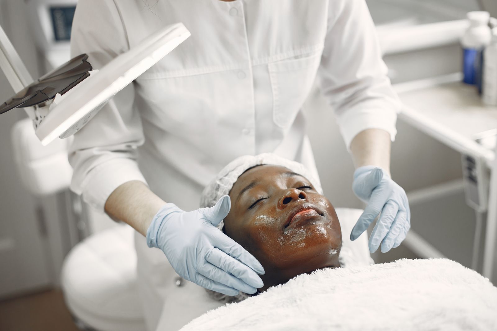 An aesthetician brushing a gold-flecked treatment mask onto the face of a woman with deep brown skin