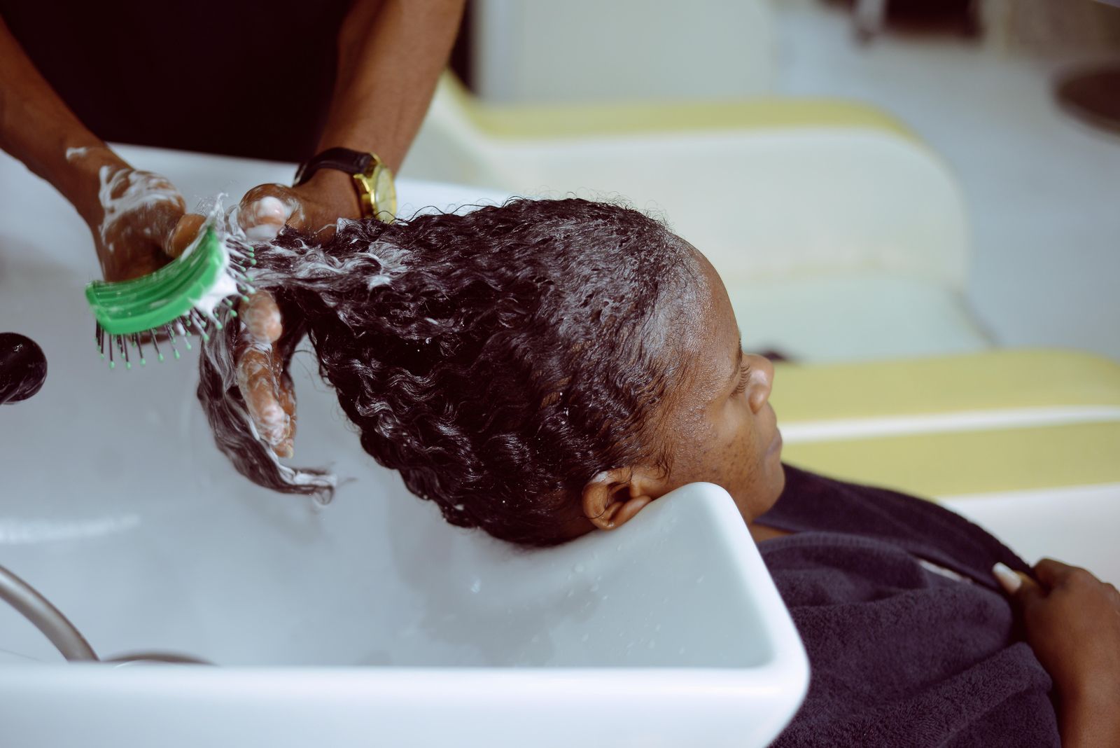 A stylist massaging a scalp treatment into a reclined Black client's hair at the basin